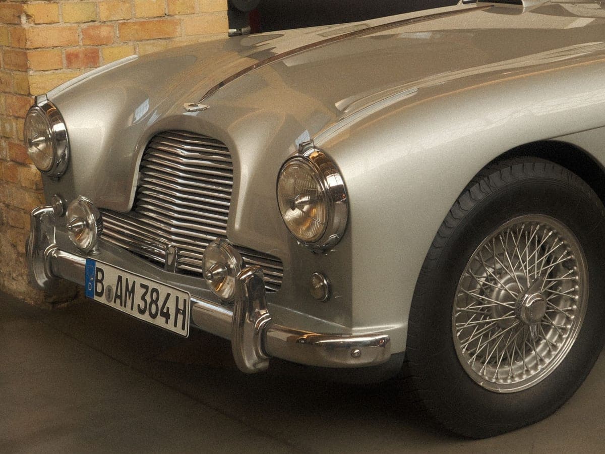 Front three-quarter view of a silver classic Aston Martin showing the grille, twin headlamps, chrome overriders and wire wheel
